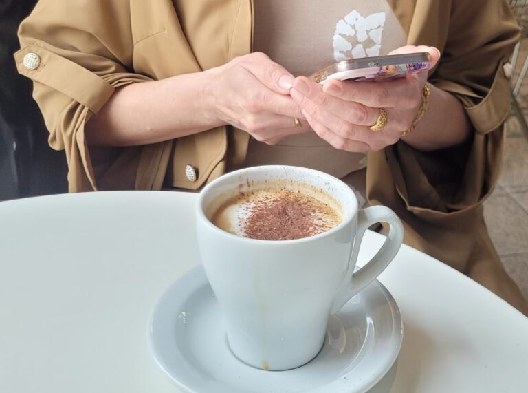 5 Warming Autumn Drinks - cozy scene with a woman in light coffee-colored clothes enjoying a cup of hot chocolate drink and reading on her phone